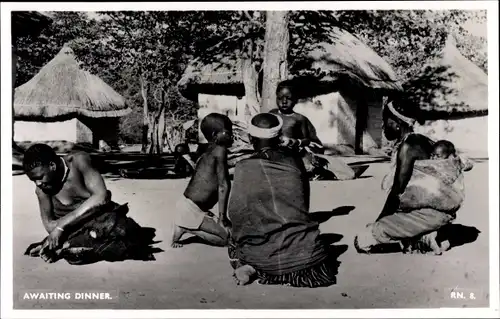 Ak Awaiting Dinner, Afrikanische Familie vor einer Hütte, Dorfansicht