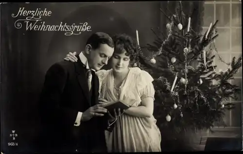 Ak Frohe Weihnachten, Paar mit Buch vor dem Tannenbaum