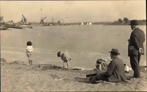 Foto Ak Familie am Strand, Männer, spielende Kinder, Hafenanlagen im Hintergrund