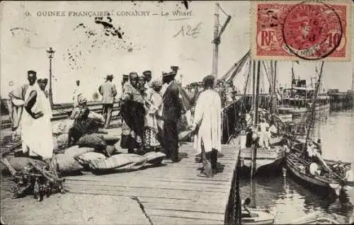 Ak Conakry Guinea, Zugangsbereich mit Personen, Waren auf dem Pier, Boote im Wasser