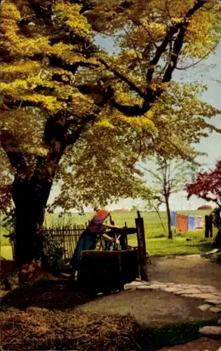 Ak Photzochromie, Frau steht an einem Brunnen, dahinter ein Haus, Bäume und ein Zaun.