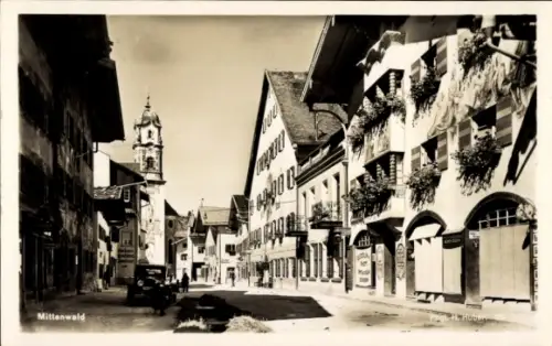 Ak Mittenwald in Oberbayern,  Straße mit Häusern, Uhrturm, Auto, Blumen, keine Personen