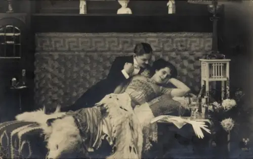 Ak Liebespaar, Mann im Smoking und Frau in einem eleganten Kleid auf einem Bett