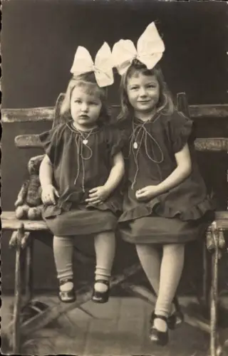 Foto Ak Kinderportrait, zwei Mädchen auf einer Holzbank, Schwestern, Haarschleifen, Teddybär