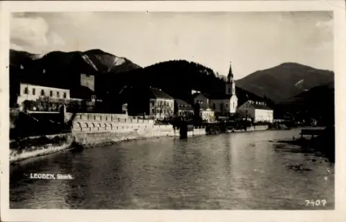 Ak Leoben Steiermark, Fluss mit Ufer, Gebäudereihe am Wasser, schmaler Kirchturm, Berge im Hinter