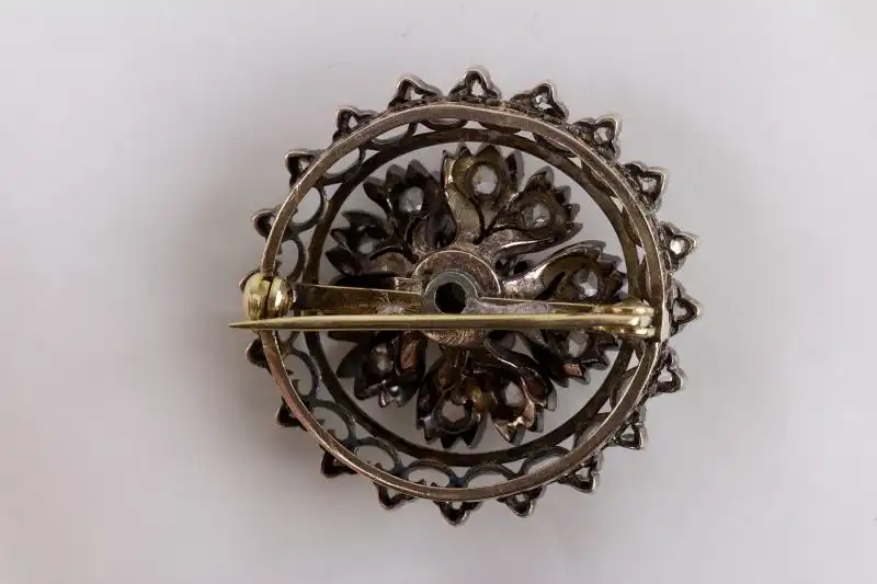 Brosche, Mitte 19. Jh., Gold mit Silber aufgelegt, verziert mit Brillanten in Altschliff, in Mitte grüner Stein, wohl später, 4 kleine Brillanten fehlen.
D: 3 cm, Gewicht: 11 g 1