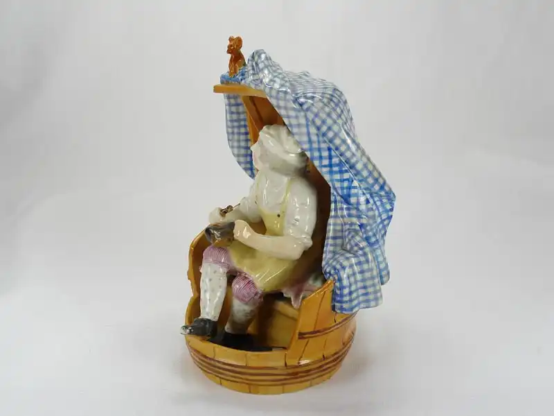 Porzellanfigur, 18./19. Jh., wohl England, Meißenkopiemarke, auf Glasur, in Fass sitzender Schuster bei der Reparatur eines Stiefels, polychrom bemalt, 2 große Brandrisse, 3 kleine Brandrisse im Fassboden, Gebrauchsspuren, sehr originell. H: 18 cm,... 1