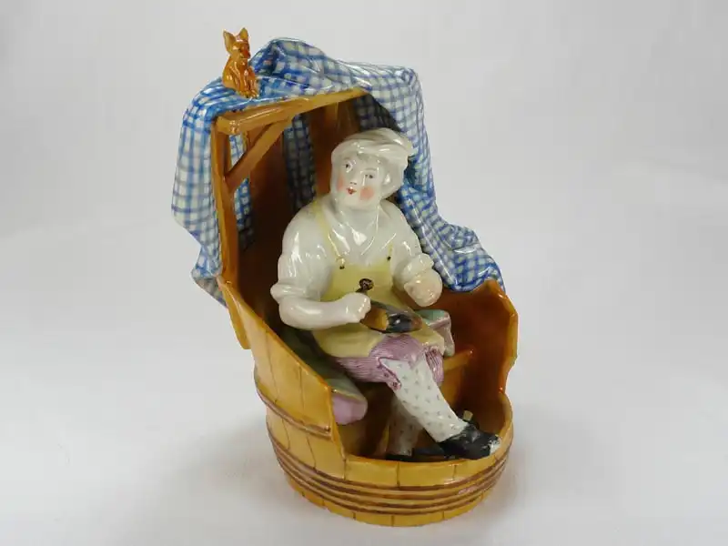 Porzellanfigur, 18./19. Jh., wohl England, Meißenkopiemarke, auf Glasur, in Fass sitzender Schuster bei der Reparatur eines Stiefels, polychrom bemalt, 2 große Brandrisse, 3 kleine Brandrisse im Fassboden, Gebrauchsspuren, sehr originell. H: 18 cm,... 2