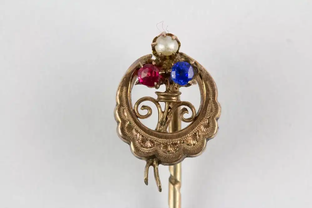 Krawattennadel, Ende 19. Jh., 585er Gold, und Silber, ungemarkt, halbmondförmig, graviert mittig Kleeblatt, mit Rubin, Perle und Saphir besetzt. L: 6 cm. 0