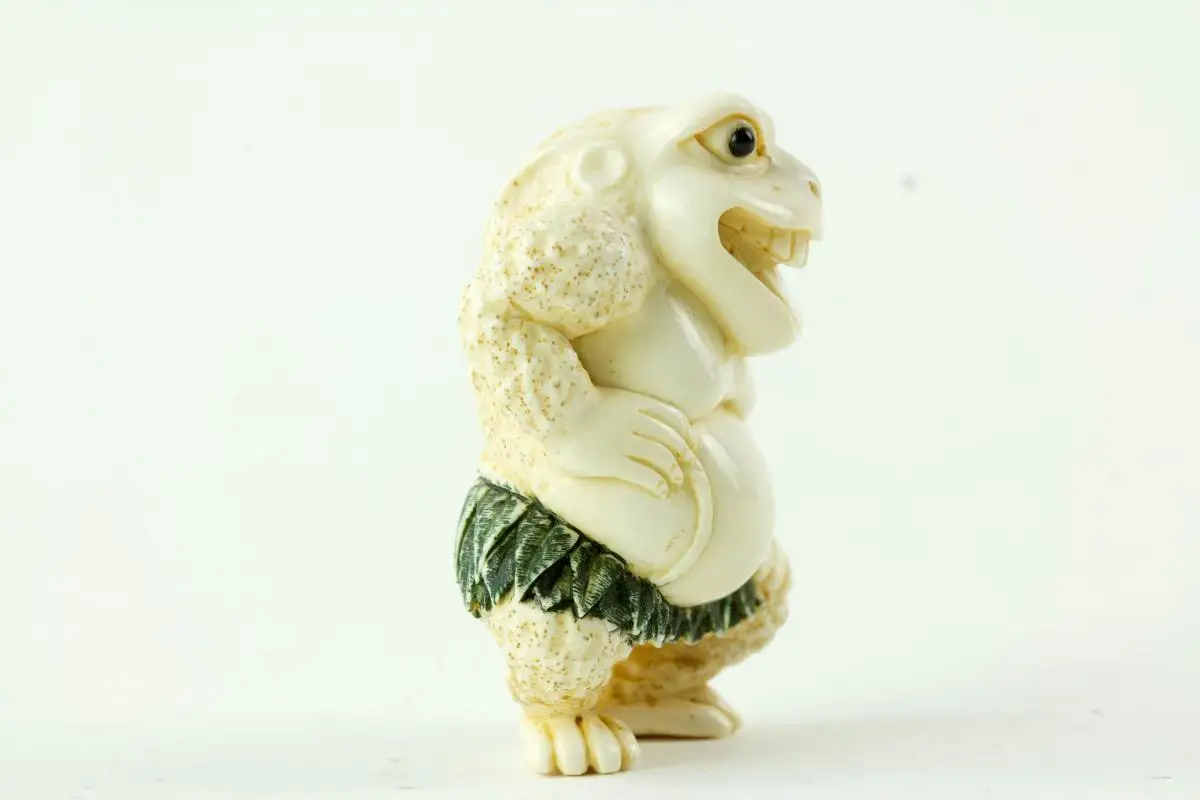 Netsuke, Japan, Taisho-Zeit, Elfenbein, signiert, humoristische Darstellung einer Kröte mit grün gefasstem Blätterrock, sehr feine Ausarbeitung, H: 4,5 cm. 1