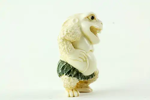 Netsuke, Japan, Taisho-Zeit, Elfenbein, signiert, humoristische Darstellung einer Kröte mit grün gefasstem Blätterrock, sehr feine Ausarbeitung, H: 4,5 cm.