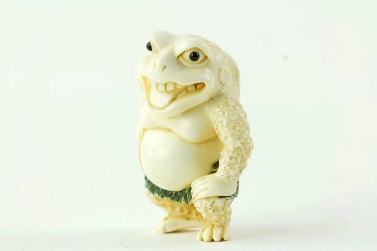 Netsuke, Japan, Taisho-Zeit, Elfenbein, signiert, humoristische Darstellung einer Kröte mit grün gefasstem Blätterrock, sehr feine Ausarbeitung, H: 4,5 cm. 3
