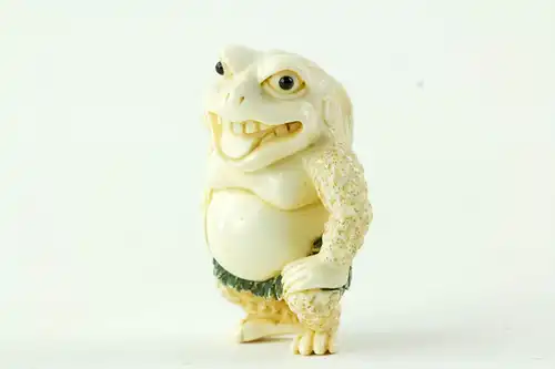 Netsuke, Japan, Taisho-Zeit, Elfenbein, signiert, humoristische Darstellung einer Kröte mit grün gefasstem Blätterrock, sehr feine Ausarbeitung, H: 4,5 cm.