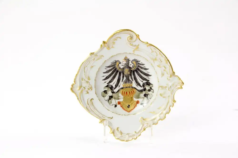 Kleiner Teller, um 1900, gemarkt KPM Berlin, blaue Zeptermarke und roter Reichsapfel, geschwungene Rokokoform, Fahne mit feiner Goldmalerei verziert, im Fond goldener Ritterhelm unter bekrönten, preußischen Adler, auf der Brust FR, handbemalt, wohl... 0