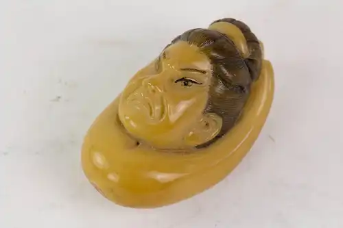 Netsuke, Japan, Meiji Zeit, um 1900, Steinnuss (Taguanuss), Guanyin Kopf in einer Hand, fein geschnitzt und braun gefasst, Gebrauchsspuren. L: 4 cm.