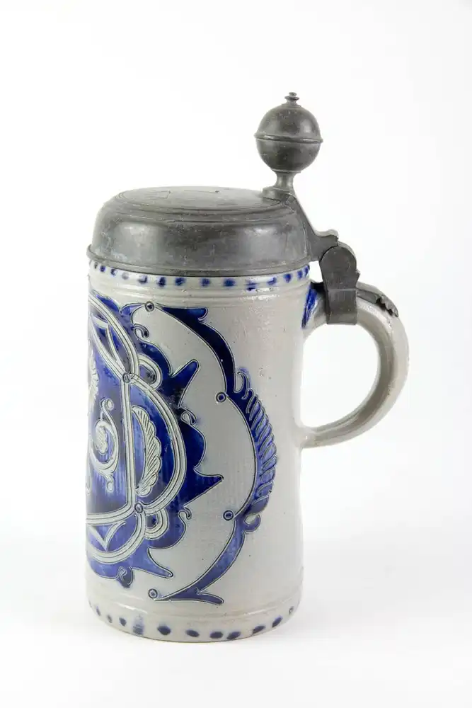 Krug, Westerwald, 19. Jh., mit Zinndeckel montiert, grauer Krug mit Blaumalerei in Ritzdekor, seitlicher Spannungsriss, sonst unbeschädigt. H: 25 cm. 2