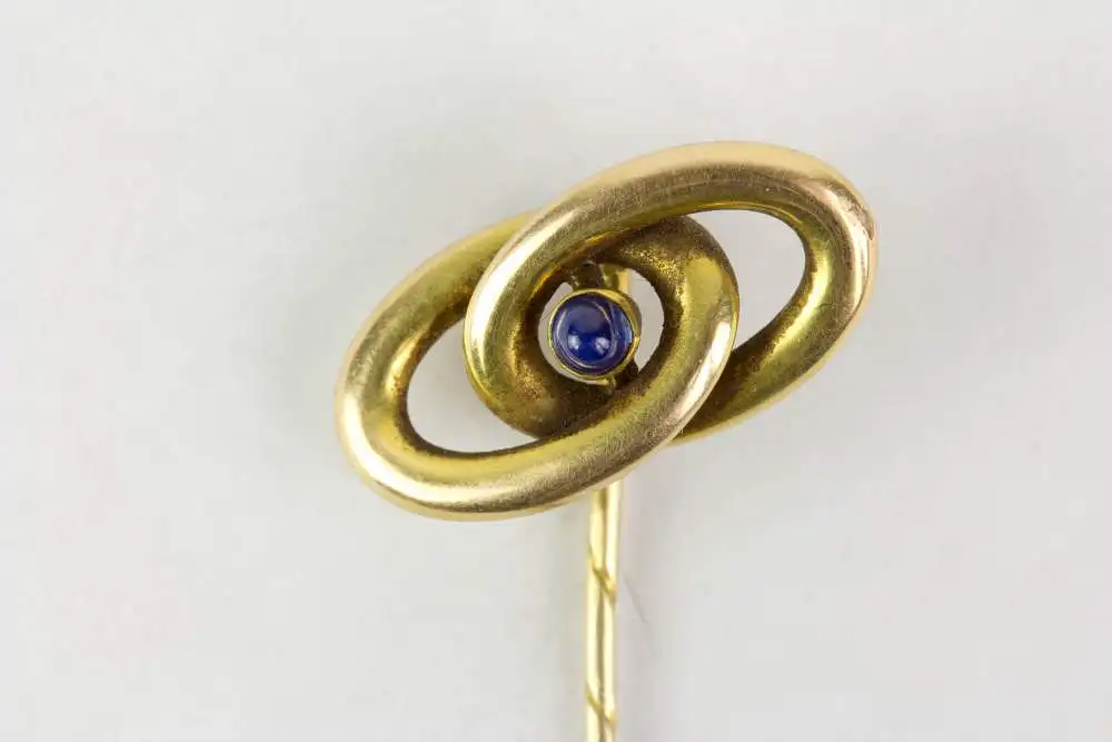 Krawattennadel, Anf. 20. Jh., gemarkt, 585er Gelbgold, zwei verschlungene Ringe, in der Mitte im Cabochon-Schliff gefasster Saphir, Gebrauchsspuren. L: 6,4 cm, 2,0 g. 0