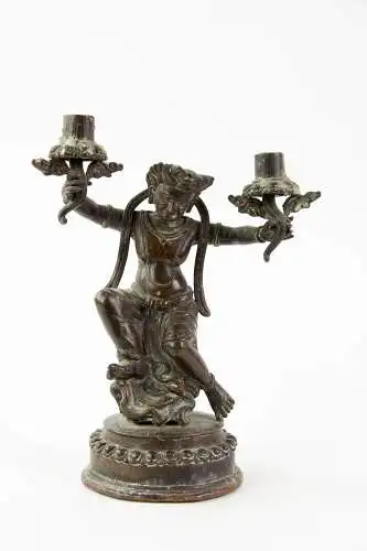 Kerzenständer - Asien, 19. Jh., zweiflammig, in Form einer asiatischen Gottheit, die in den Händen zwei blumenförmige Tüllen hält, Sockel mit Resten einer roten Bemalung, Gebrauchsspuren, H 21 cm.