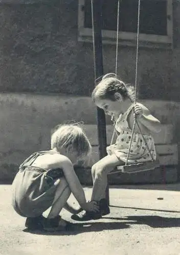 Postcard children types playing at the swing young boy tying young girl shoelaces