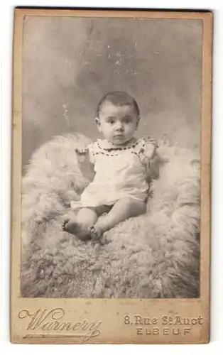 Fotografie Warnery, Elbeuf, Baby im weissen Kleid auf Felldecke sitzend