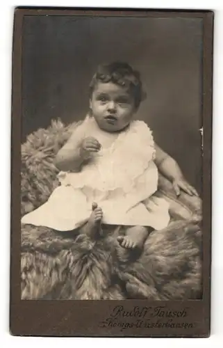 Fotografie Rudolf Tausch, Königs-Wusterhausen, niedliches kleines Mädchen im weissen Kleid auf Felldecke sitzend
