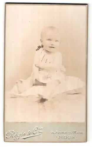 Fotografie Regis & Cie, Paris, lächelndes kleines Mädchen mit blondem Haar im weissen Kleidchen mit schwarzer Schleife