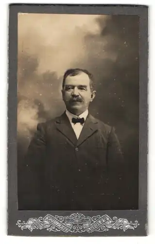 Fotografie F. de Leeuw, Salies-de-Bearn, Portrait Herr im Anzug mit Fliege vor Wolkenkulisse