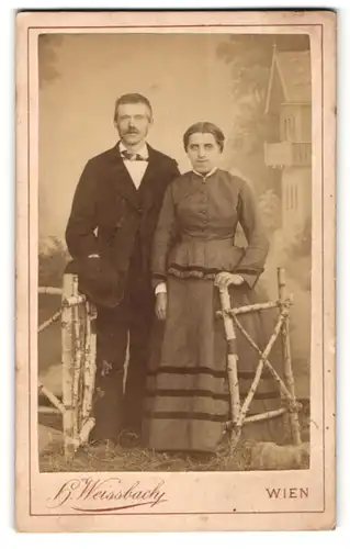 Fotografie H. Weissbach, Wien, hübsches Paar in edler Kleidung am Holzzaun stehend