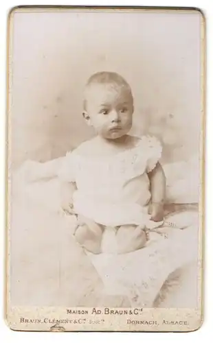 Fotografie Braun, Dornach Alsace, Kleines Baby in weissem Kleidchen schaut verdutzt in Richtung Kamera