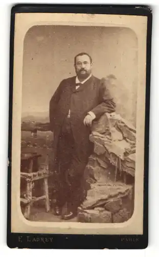 Fotografie E. Ladrey, Paris, charmanter korpulenter Herr im eleganten Mantel am Felsen stehend