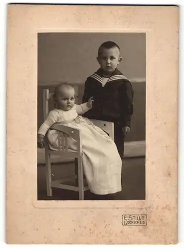 Fotografie E. Stille, Lüdenscheid, Kleiner Junge steht neben Baby auf Stuhl