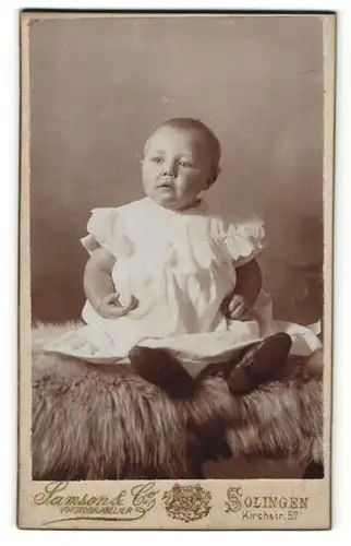 Fotografie Samson & Co, Solingen, Baby in Rüschenhemdchen sitzt auf Felldecke
