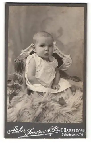 Fotografie Atelier Samson & Co, Düsseldorf, niedliches Baby im weissen Kleidchen auf Felldecke sitzend