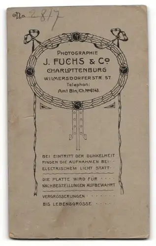 Fotografie J. Fuchs & Co, Berlin, niedlich blickendes Kleinkind im weissen Kleidchen auf Felldecke sitzend