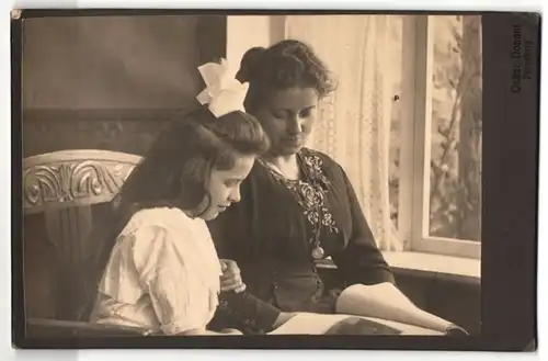 Fotografie Oskar Donant, Pinneberg, hübsche junge Mutter mit lächelnder Tochter und Haarschleife in Zeitschrift schauend