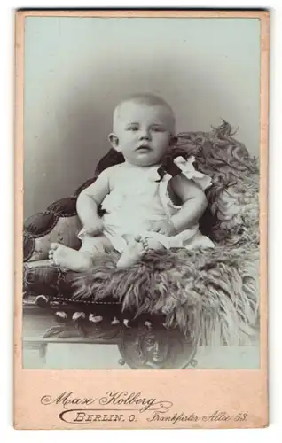 Fotografie Max Kolberg, Berlin, zuckersüsses Baby mit blondem Haar, Kleidchen und nackten Füssen auf einer Felldecke