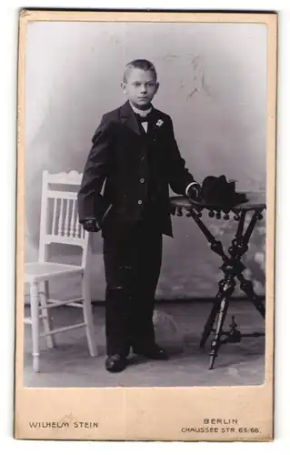 Fotografie Wilhelm Stein, Berlin, Junge im Anzug und Hut an Tisch stehend