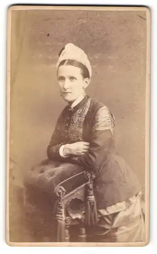 Fotografie Atelier W. Runicles & Sons, Windsor, stehende Frau mit dunkler Jacke und hellem Hut auf Hocker gelehnt