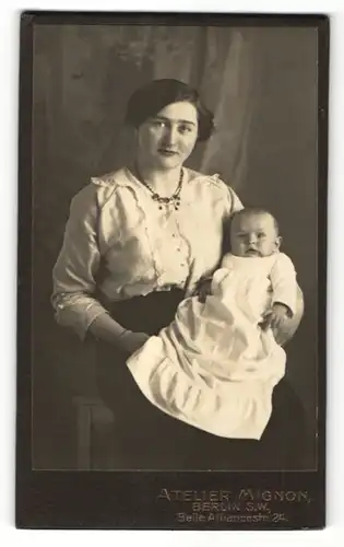 Fotografie Atelier Mignon, Berlin, strahlende junge Mutter mit Halskette hält süsses Baby im Kleidchen im Arm