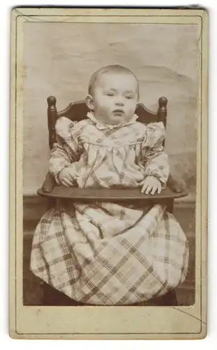 Fotografie Fotograf und Ort unbekannt, Kleinkind mit kariertem Kleid im Kinderstuhl