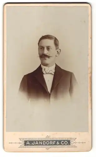 Fotografie A. Jandorf & Co., Berlin, Portrait charmanter dunkelhaariger Mann mit Schnurrbart und Fliege im Jackett