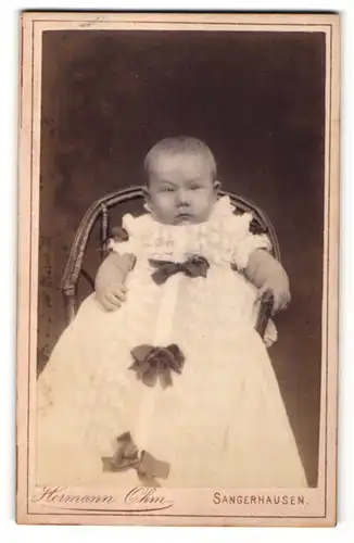 Fotografie Hermann Ohm, Sangerhausen, Portrait Baby im weissen Kleid mit Schleifchen auf einem Stuhl