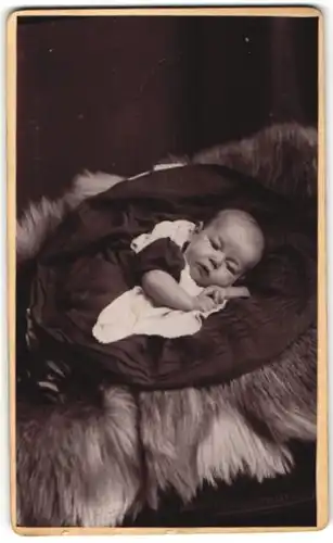 Fotografie Garralt, Dewsbury, zuckersüsses Baby im hübschen Kleidchen auf Felldecke liegend