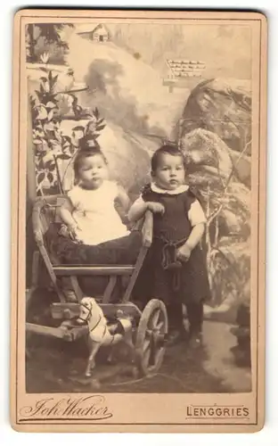 Fotografie Joh. Wacker, Lenggries, Portrait zweier Kleinkinder in bürgerlicher Kleidung im Karren mit Spielzeugpflerd