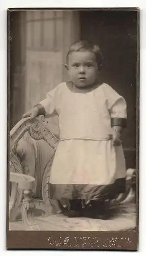 Fotografie Fotograf und Ort unbekannt, Portrait Kleinkind im weissen Kleid auf einer Bank