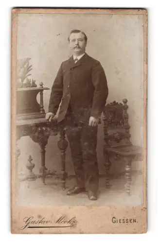 Fotografie Gustav Mook, Giessen, Portrait älterer Herr mit Zwirbelbart im Anzug an Tisch gelehnt
