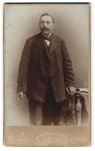 Fotografie J. Fuchs, Berlin, Portrait älterer bürgerlicher Herr mit Fliege im Anzug an Tisch gelehnt