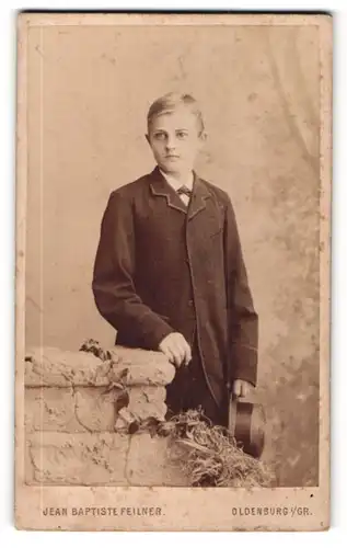 Fotografie Jean Baptiste Feilner, Oldenburg i / Gr., Portrait hübscher Knabe im Mantel mit Hut an Sockel gelehnt