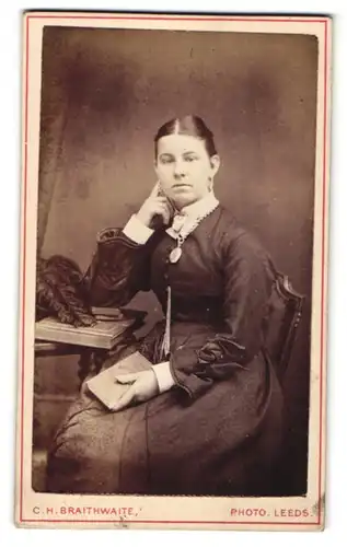Fotografie C. H. Braithwaite, Leeds, Portrait, Portrait junge Dame mit Amulett u. Buch am Tisch sitzend