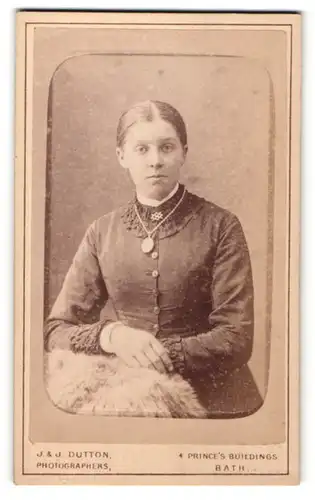 Fotografie J. & J. Dutton, Bath, Portrait junge Dame mit Amulett u. zurückgebundenem Haar an Fell gelehnt
